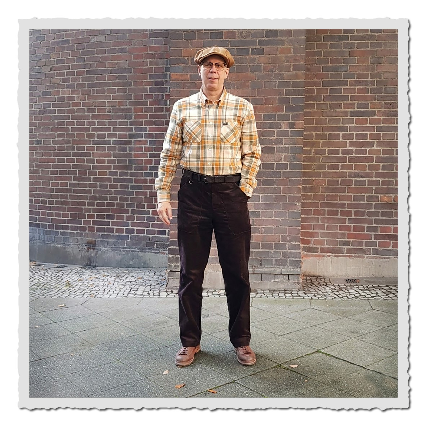 Person steht vor Backsteinfassade, Stadtstraße mit Pflaster und trägt schwarzbraune Cordhose und Karohemd.